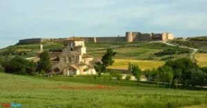 
Vista general de Urueña que, situada en un altozano, es conocida por 'El Mirador de la Tierra de Campos'. Su muralla, de los siglos XII y XIII, rodea la ciudad y tiene dos puertas, mirando al norte, la puerta principal 'Puerta del Azogue' y al sur el 'Arco de la Villa'. A la derecha de la muralla, su castillo, en el que vivieron personajes importantes de la historia de España. En primer plano y, fuera de la villa amurallada, la ermita de 'Nuestra Señora de la Anunciada' que, de estilo románico lombardo con su cimborrio octogonal, fue construida en el siglo XI.